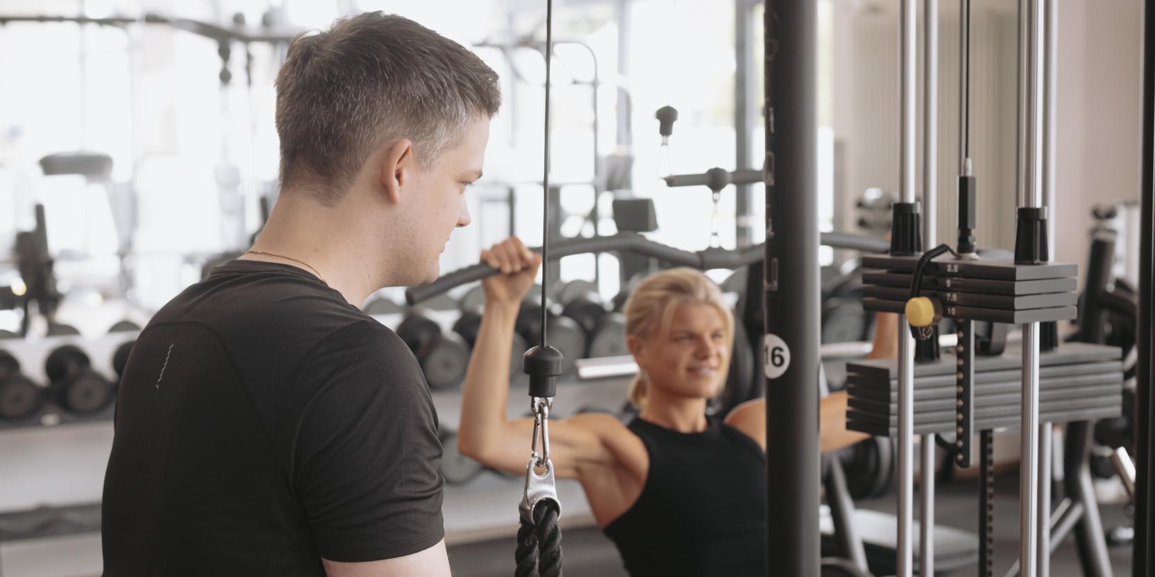 Zwei Personen im Fitnessstudio nutzen ein Kabelzuggerät zum Training. Der Mann steht vor dem Gerät, die Frau sitzt dahinter.