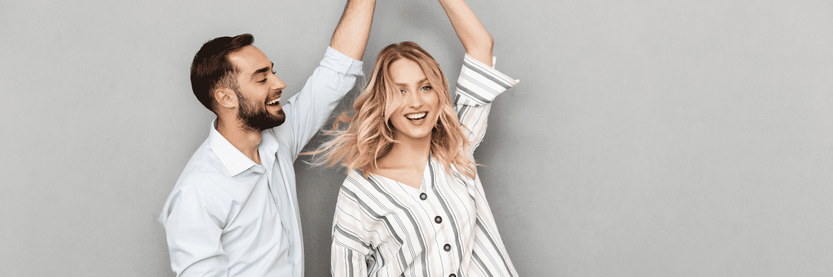 Ein Mann und eine Frau stehen vor einer grauen Wand, lächeln und halten ihre Hände über ihren Köpfen.