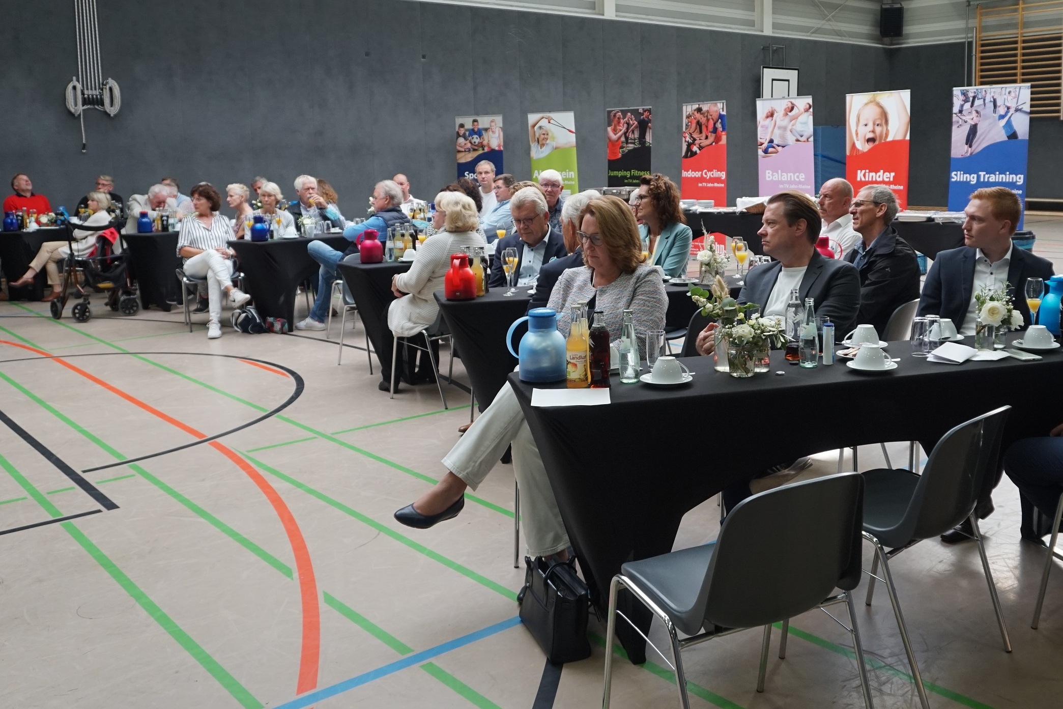 Veranstaltung in einer Sporthalle: Teilnehmer sitzen an Tischen mit Getr&auml;nken und Werbebannern im Hintergrund.