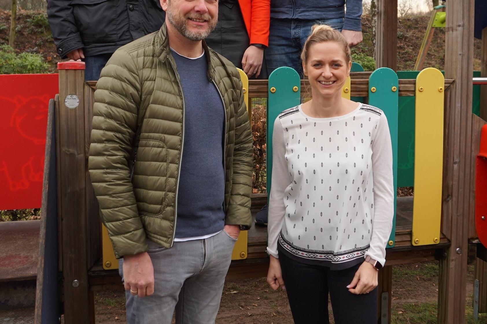 F&uuml;nf Personen stehen auf einem Spielplatz, umgeben von bunten Spielger&auml;ten und B&auml;umen im Hintergrund.