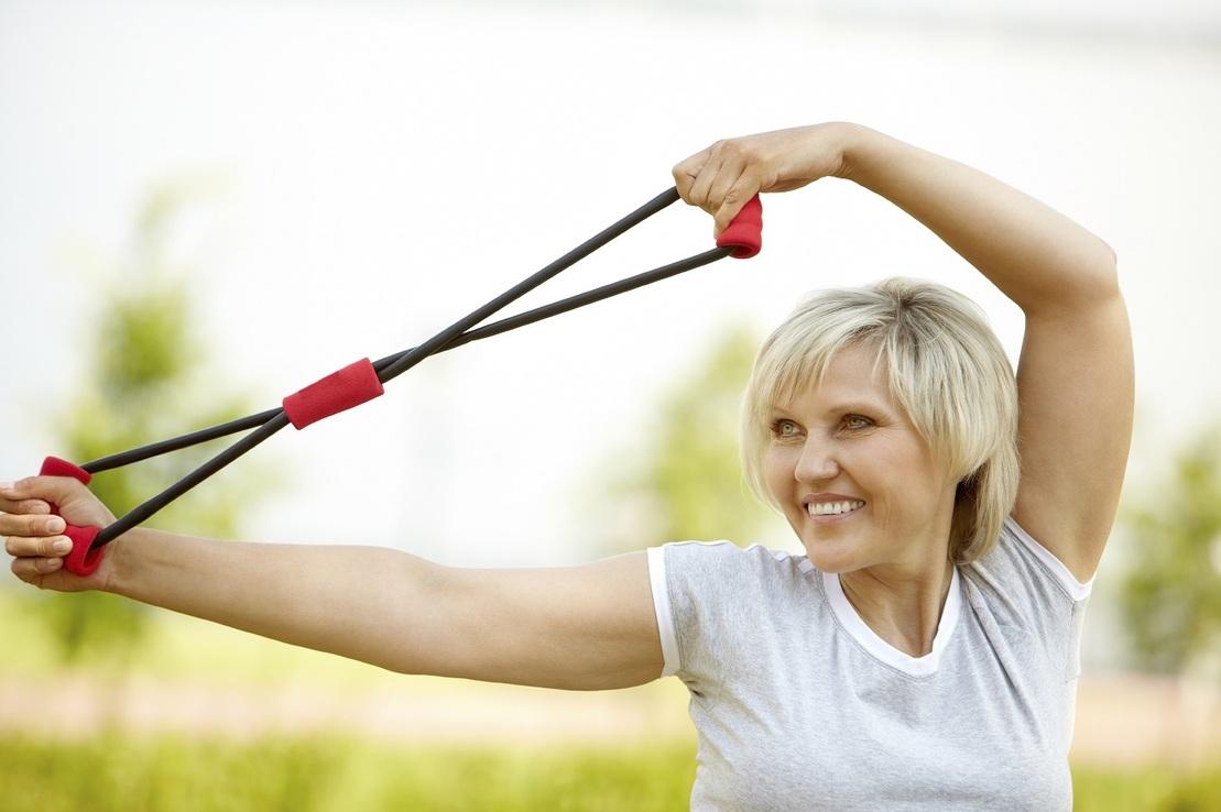&Auml;ltere Frau dehnt ein Fitnessband im Freien und l&auml;chelt dabei, umgeben von gr&uuml;ner Natur.