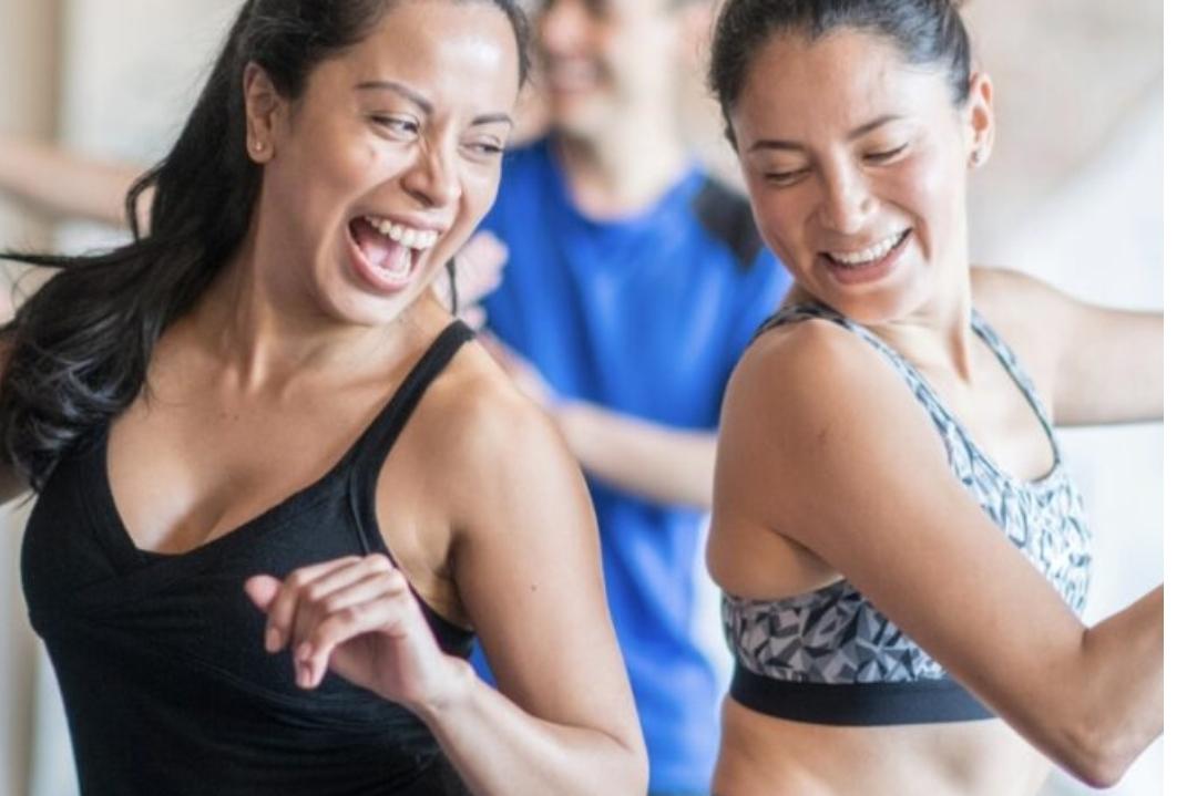 Zwei Frauen tanzen l&auml;chelnd in sportlicher Kleidung, w&auml;hrend ein Mann im Hintergrund ebenfalls fr&ouml;hlich mitmacht.
