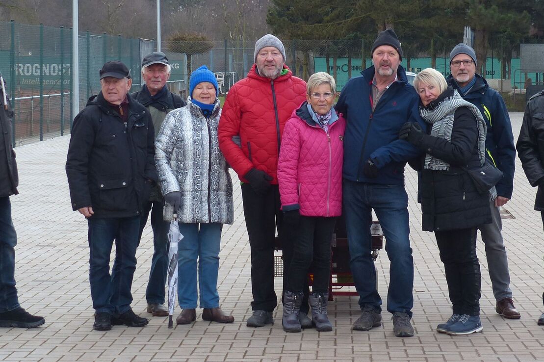 Gruppe von zehn Personen auf einem Platz, umgeben von Tennispl&auml;tzen, an einem k&uuml;hlen, bew&ouml;lkten Tag.