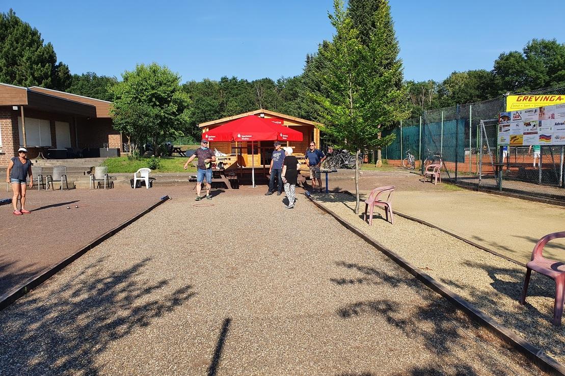 Schotterplatz mit G&auml;sten, Holzst&auml;nden und einem roten Pavillon, umgeben von B&auml;umen und Tennispl&auml;tzen.