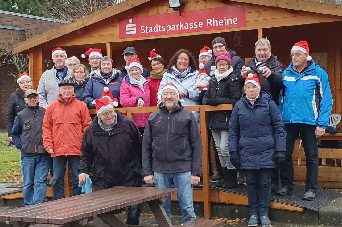 Gruppe von Menschen in Weihnachtsm&uuml;tzen vor einer Holzschutzh&uuml;tte mit Logo der Stadtsparkasse Rheine.