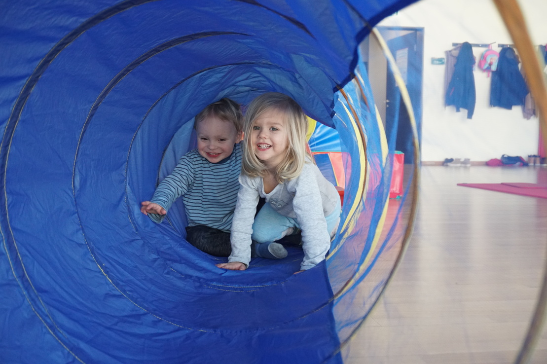 Zwei Kinder kriechen l&auml;chelnd durch einen blauen Spieltunnel in einem hellen Raum.