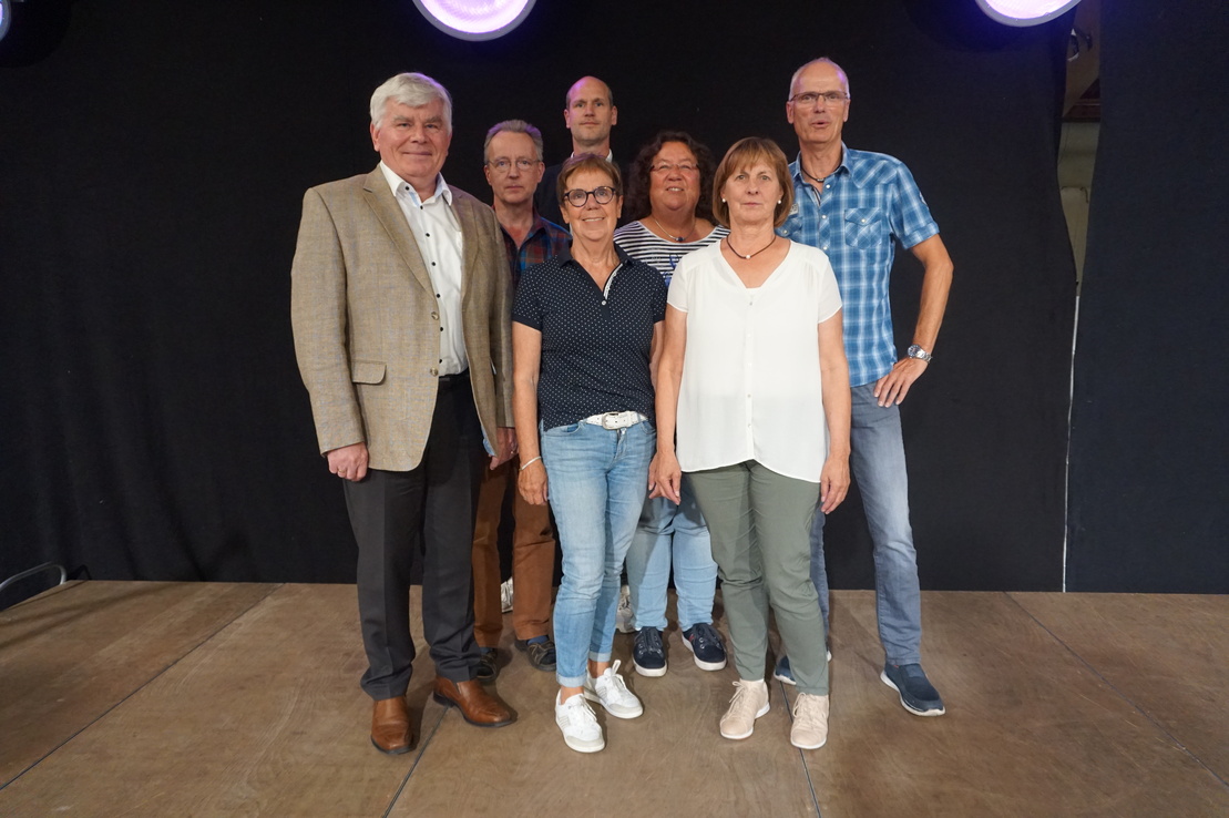 Gruppenfoto von sieben Personen auf einer B&uuml;hne mit schwarzem Hintergrund und sanfter Beleuchtung.