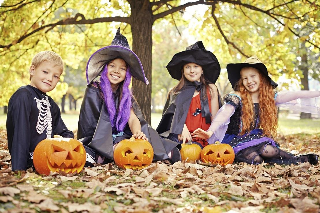 Vier Kinder in Halloween-Kost&uuml;men sitzen auf buntem Herbstlaub mit geschnitzten K&uuml;rbissen vor einem Baum.