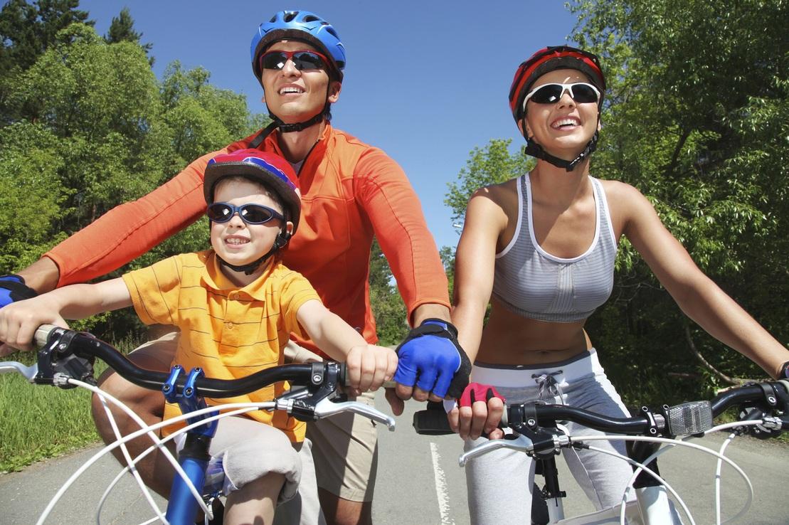 Familie beim Fahrradfahren auf einem Waldweg, l&auml;chelnd, in Radhelmen und Sonnenbrillen, bei sonnigem Wetter.
