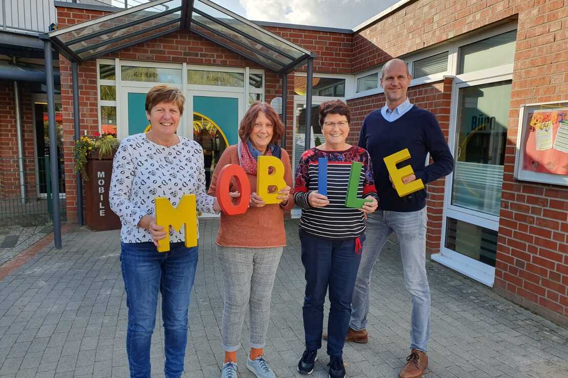 Vier Personen stehen vor einem Geb&auml;ude und halten bunte Buchstaben, die das Wort "MOBILE" bilden.
