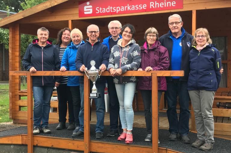 Gruppe von neun Personen mit Pokal vor einer Holzh&uuml;tte der Stadtsparkasse Rheine, umgeben von gr&uuml;nem Gel&auml;nde.