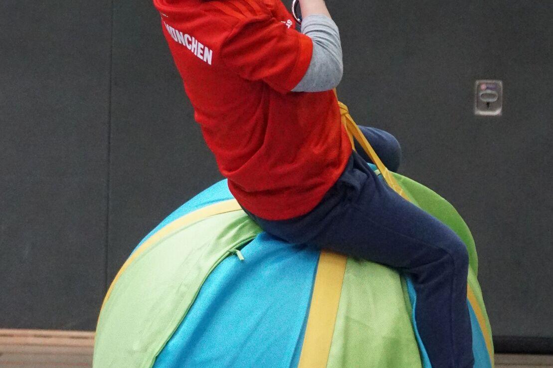 Ein l&auml;chelnder Junge in rotem T-Shirt schwingt auf einem gro&szlig;en, bunten Luftballon in einer Sporthalle.