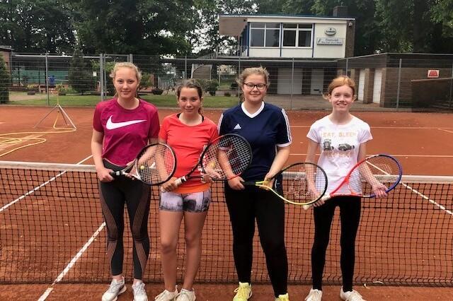 Vier Jugendliche stehen vor einem Tennisnetz, jede mit einem Schl&auml;ger in der Hand, auf einem Sandplatz.