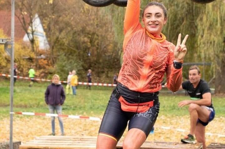 Sportlerin schwingt fr&ouml;hlich an Reifen, tr&auml;gt orangefarbene Jacke und Shorts, im Hintergrund weitere Teilnehmer und Holzpaletten.