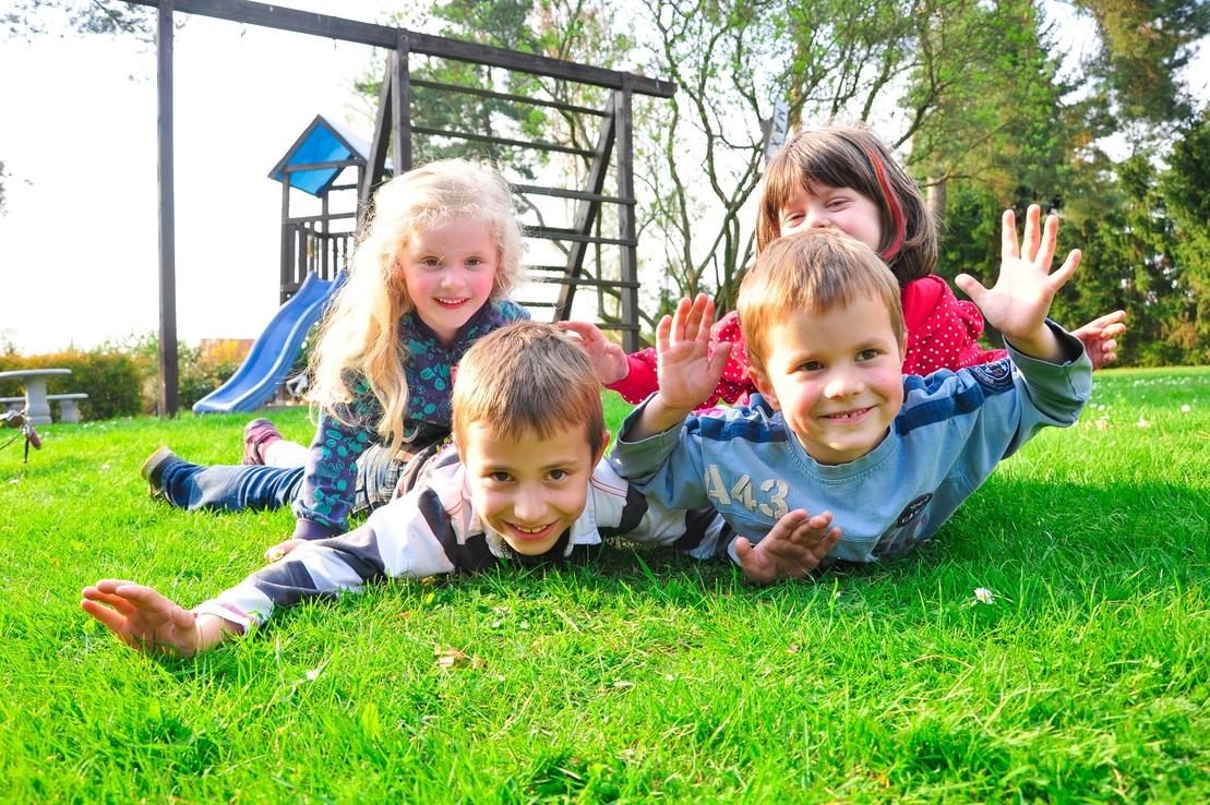 Vier Kinder liegen l&auml;chelnd auf der Wiese, im Hintergrund ein Spielplatz mit Rutsche und Kletterger&uuml;st.