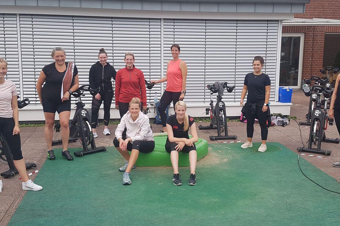 Gruppe von acht Frauen in Sportkleidung, posierend in einem Fitnessbereich mit Fahrr&auml;dern auf gr&uuml;nem Untergrund.