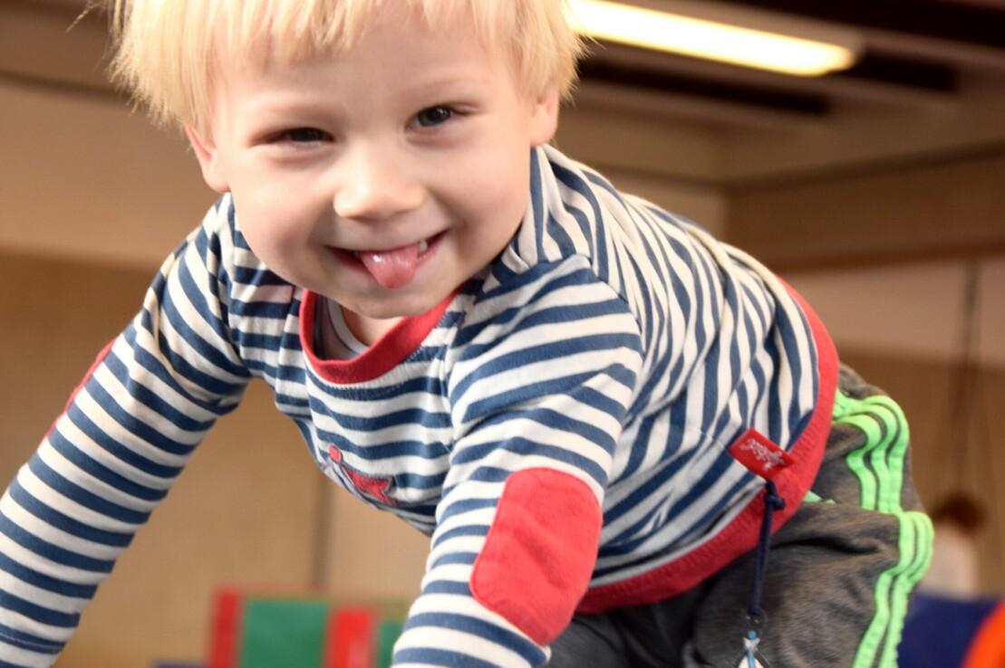 L&auml;chelnder Junge mit blonden Haaren, der auf einer Turnmatte spielt und die Zunge herausstreckt.