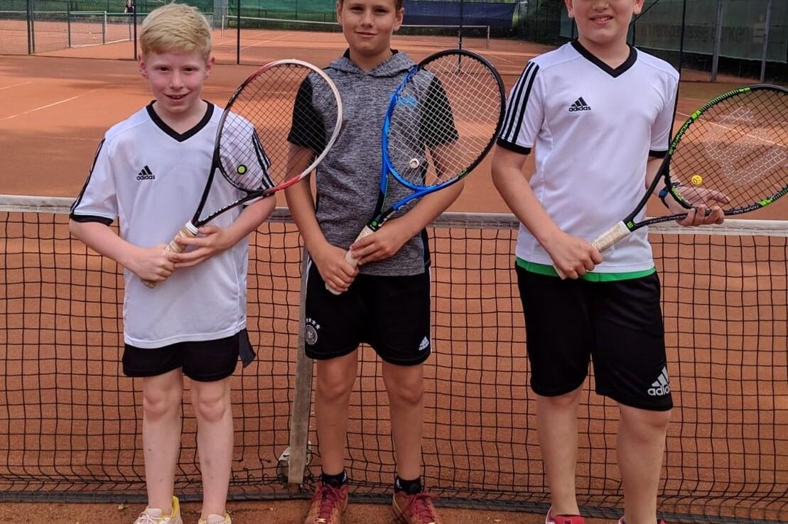 Drei junge Tennisspieler stehen am Netz eines Sandplatzes, jeder mit einem Schl&auml;ger in der Hand.