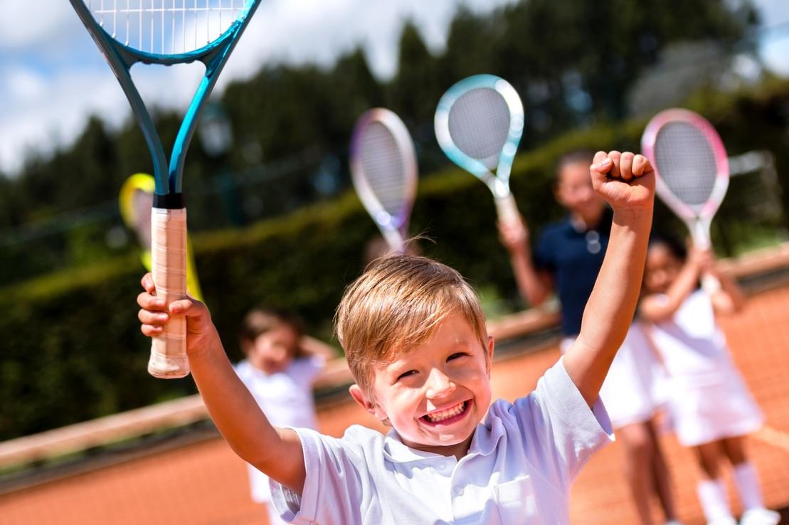 Junge mit Tennisschl&auml;ger in der Hand, jubilierend auf einem Tennisplatz, im Hintergrund weitere Kinder mit Schl&auml;gern.
