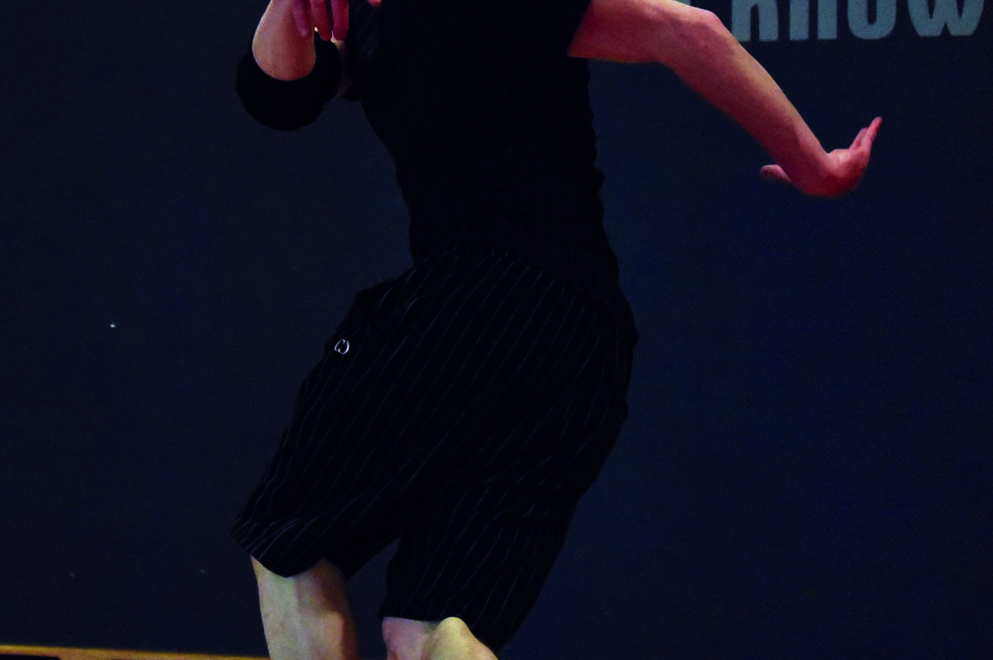 T&auml;nzer in schwarzer Kleidung und M&uuml;tze f&uuml;hrt einen Schritt auf einem Holzfu&szlig;boden aus, Hintergrund mit Schriftz&uuml;gen.