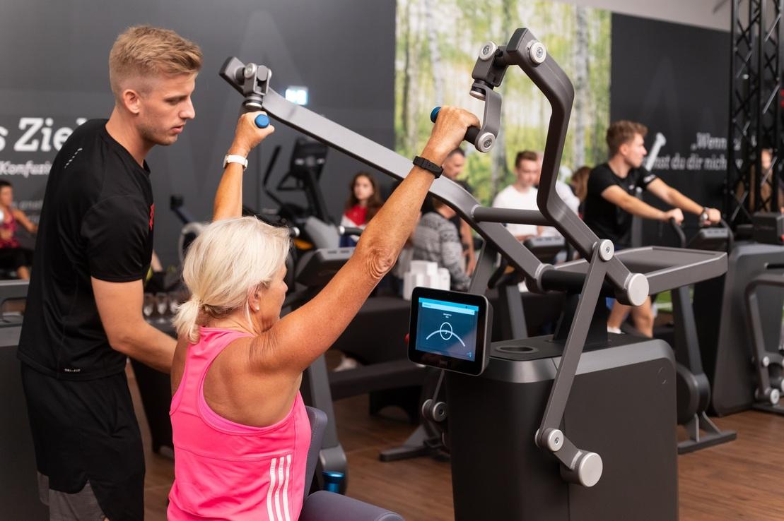 Trainer unterst&uuml;tzt &auml;ltere Frau beim Krafttraining an einem Fitnessger&auml;t in einem modernen Fitnessstudio.