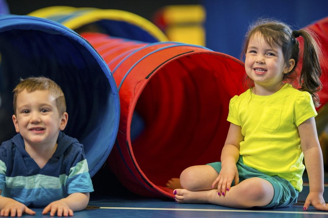 Zwei l&auml;chelnde Kinder sitzen vor bunten Spieltunneln in einer Indoor-Spielarena.