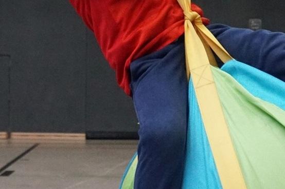 Junge mit rotem T-Shirt l&auml;chelt, w&auml;hrend er an einem bunten Schaukeltier in einer Sporthalle h&auml;ngt.