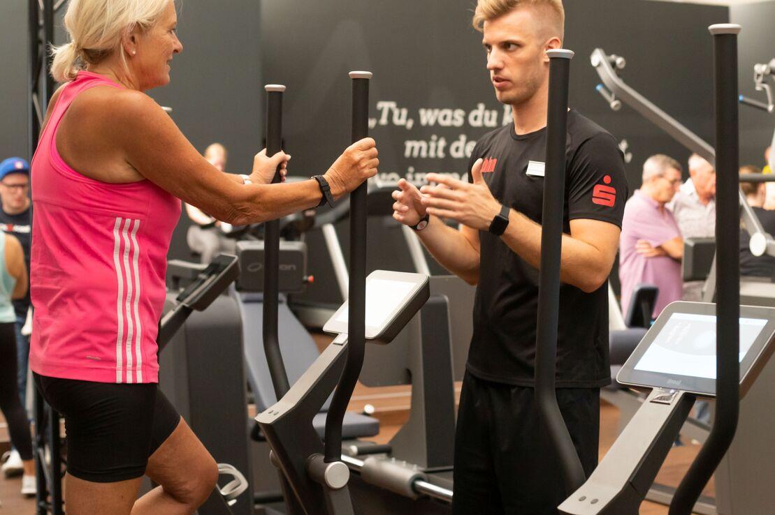 Frau auf einem Crosstrainer im Gespr&auml;ch mit einem Trainer in einem Sportstudio. Menschen im Hintergrund.