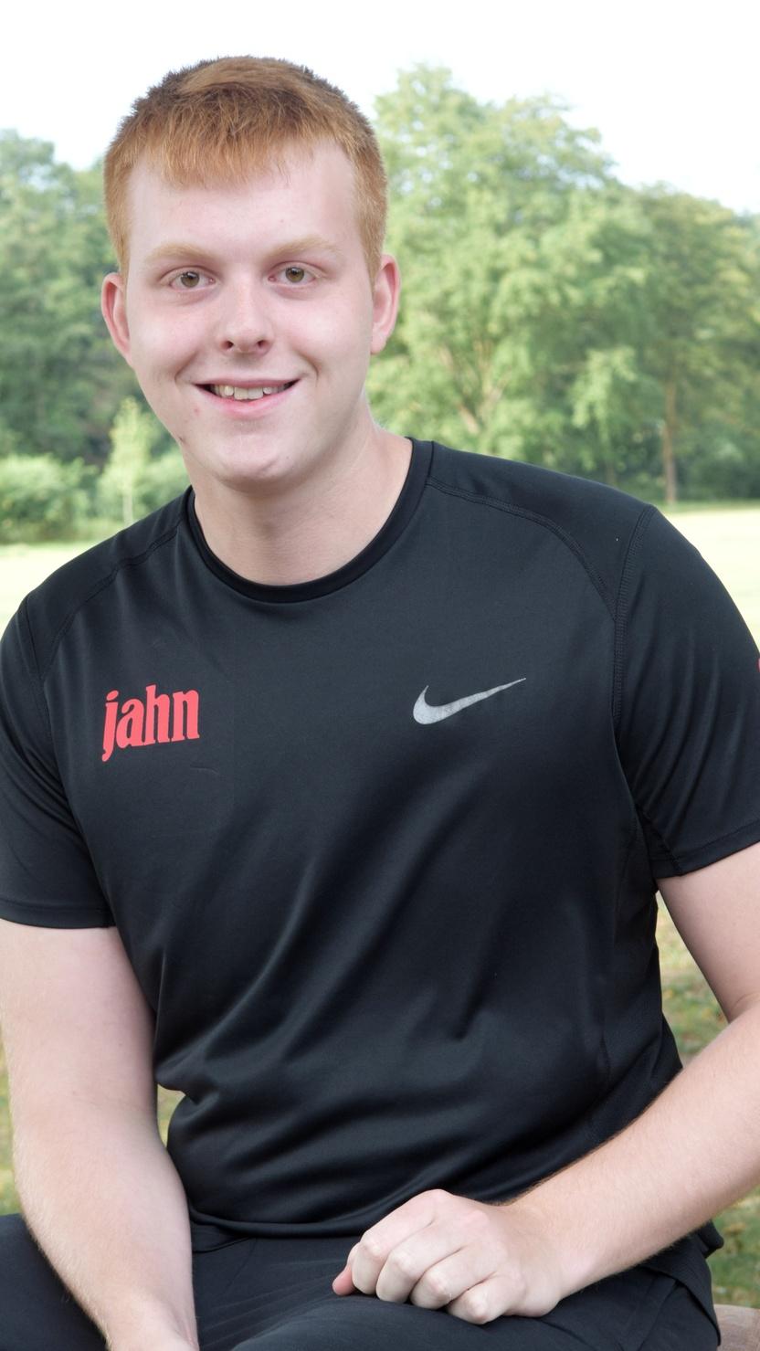 Junger Mann mit kurzen roten Haaren sitzt in einem Park, tr&auml;gt ein schwarzes Sportshirt mit roten Details.