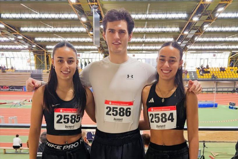 Drei Athleten in Sportkleidung mit Startnummern stehen l&auml;chelnd auf einer Trib&uuml;ne in einer sportlichen Halle.