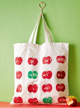 Stofftasche mit Apfelmuster in Rot und Gr&uuml;n, aufgeh&auml;ngt an einer gr&uuml;nen Wand. Ein roter Apfel liegt auf einer orangefarbenen Oberfl&auml;che.