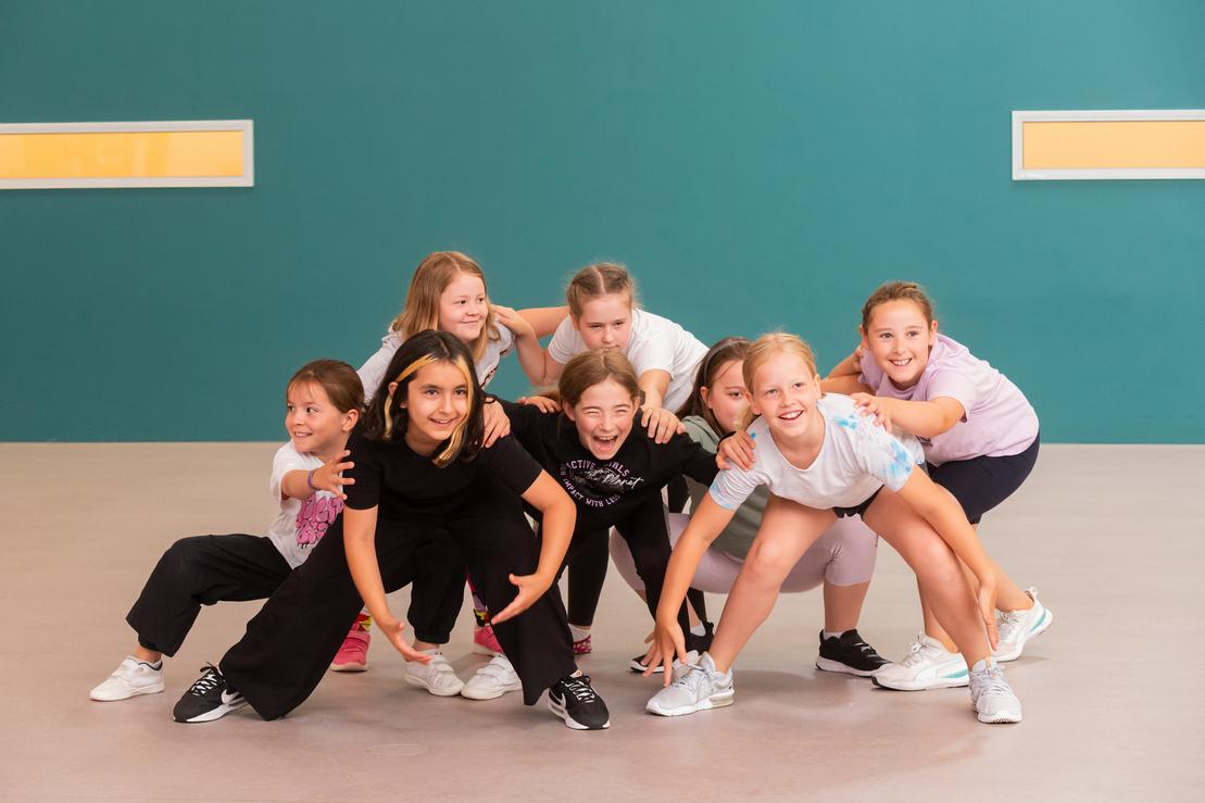 Gruppe von acht M&auml;dchen in Sportkleidung, die in einem kreativen Pose auf einem Sportboden stehen.