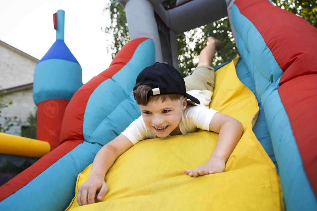 Junge rutscht lachend von einer bunten, aufblasbaren Rutsche in einem Garten.