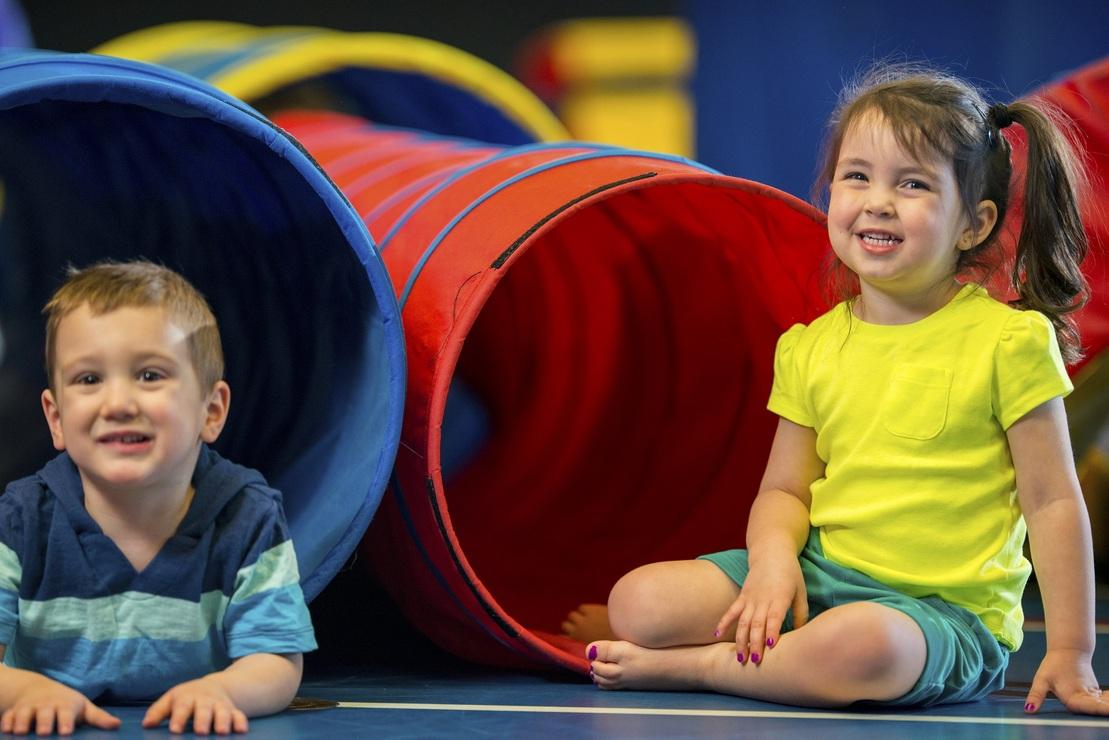 Zwei Kinder sitzen l&auml;chelnd auf einer blauen Unterlage neben bunten Tunnelr&ouml;hren in einem Spielbereich.