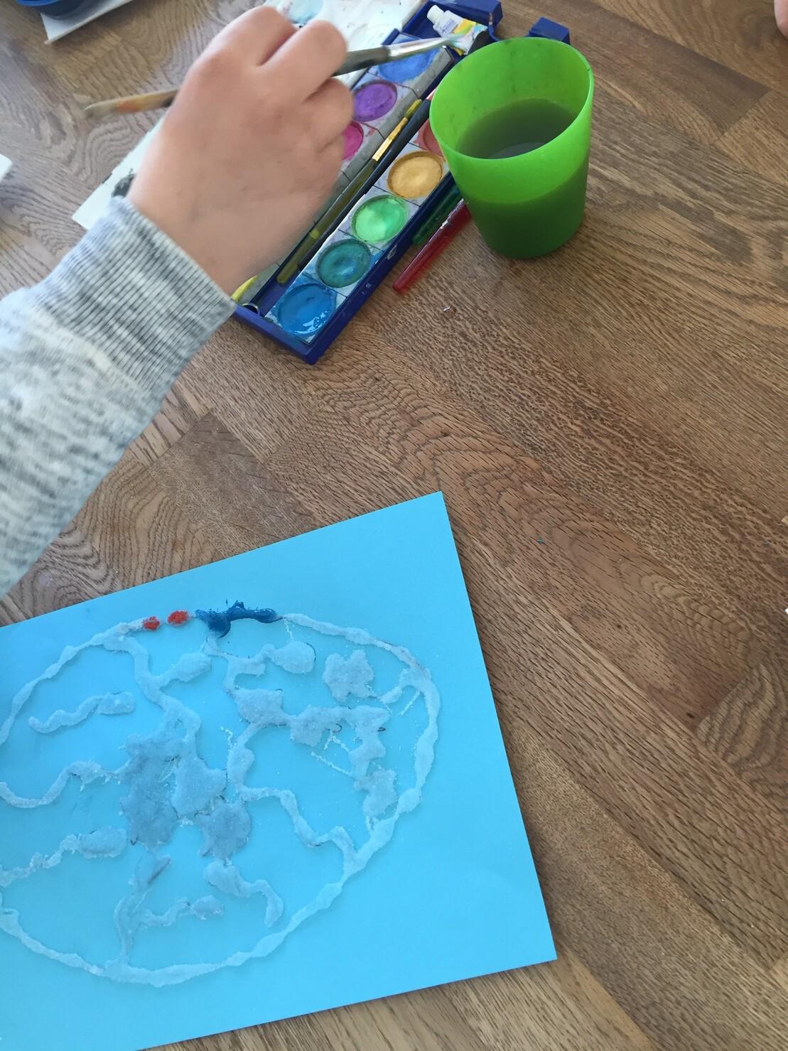 Ein Kind malt mit Wasserfarben auf blauem Papier, w&auml;hrend eine Hand einen Pinsel h&auml;lt und eine gr&uuml;ne Tasse daneben steht.