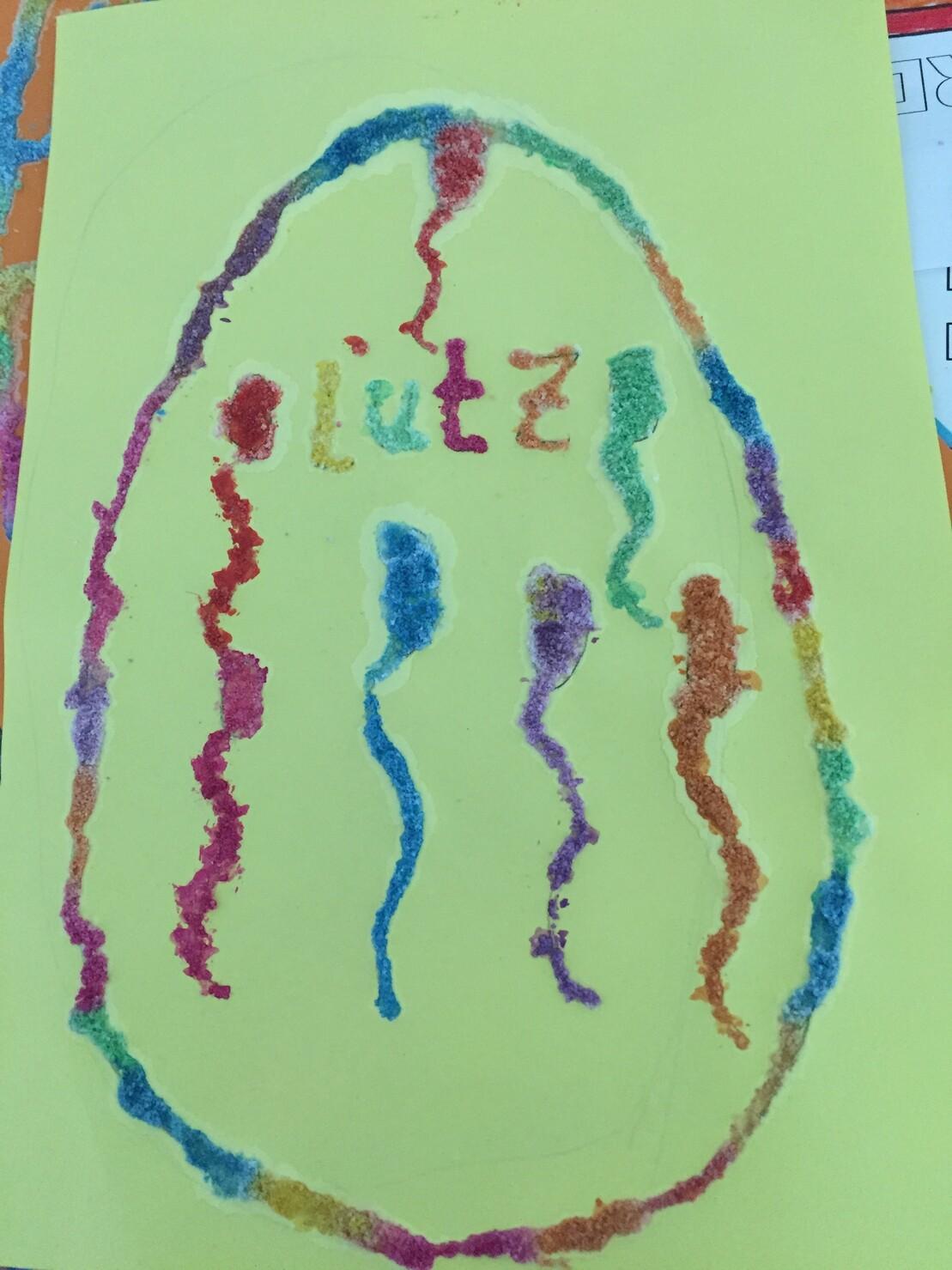 Bunte Verzierung eines Ostereis aus Sand auf gelbem Papier mit dem Wort &bdquo;l&uuml;tz&ldquo; in der Mitte.