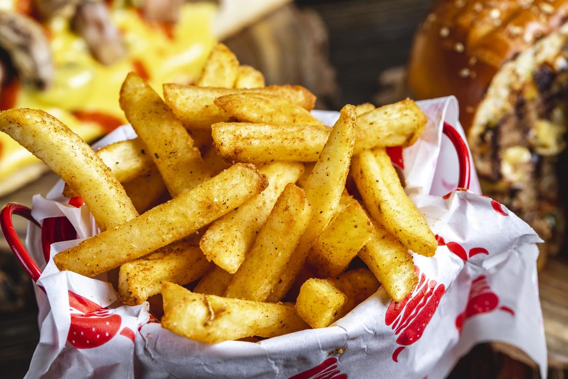 Ein Korb mit knusprigen, gew&uuml;rzten Pommes frites auf einer Holzoberfl&auml;che, im Hintergrund ein Burger.