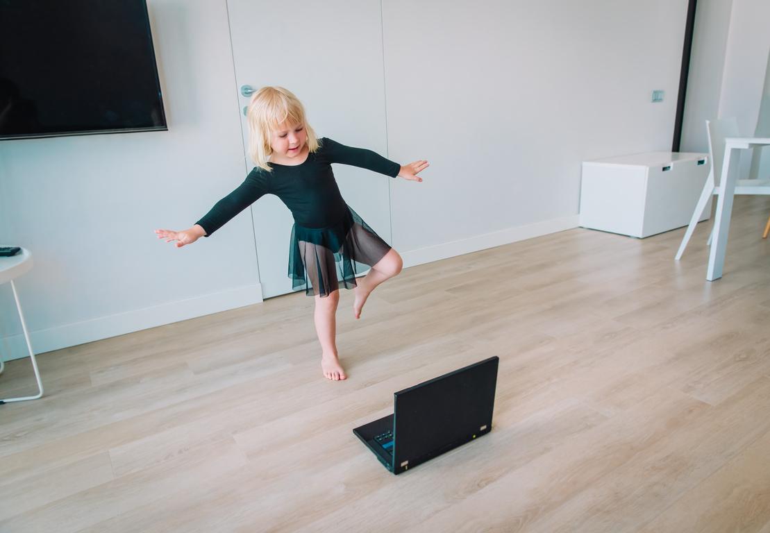 M&auml;dchen in einem Tutu tanzt barfu&szlig; vor einem Laptop in einem modernen Raum mit Holzfu&szlig;boden.