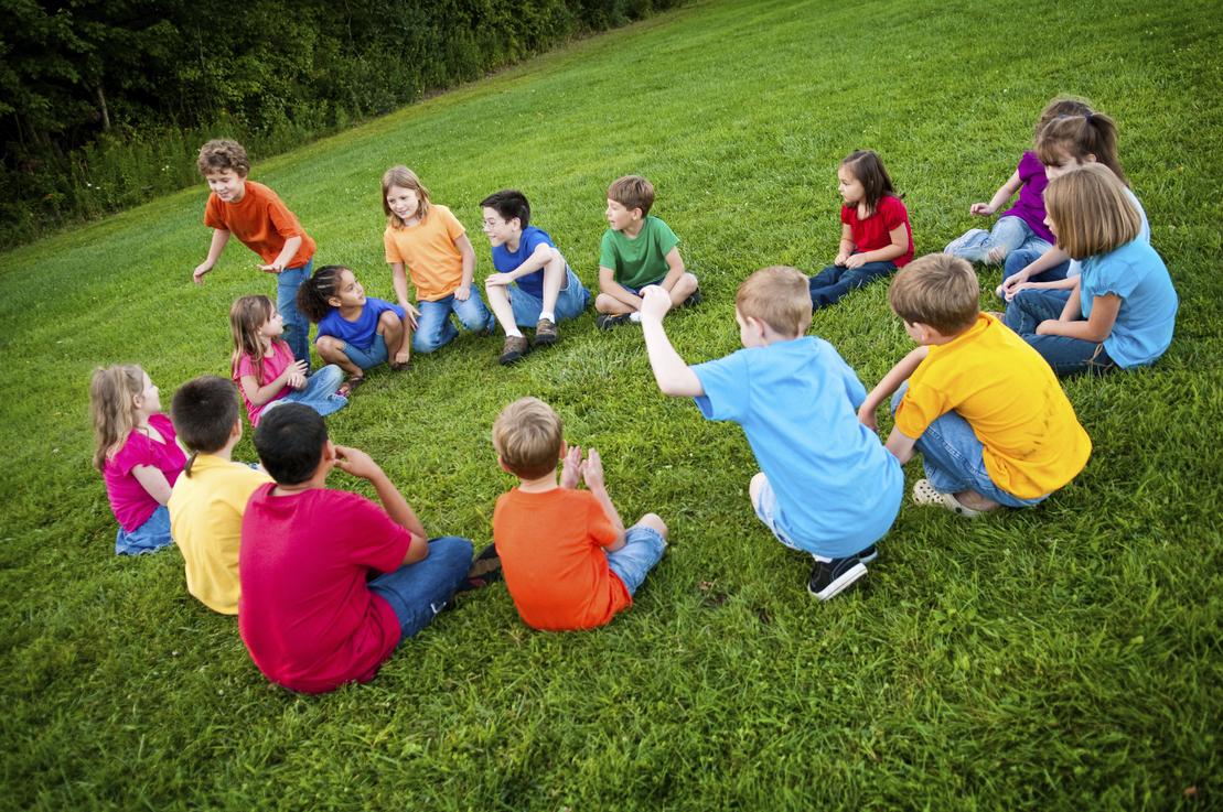 Gruppe von Kindern in bunten T-Shirts sitzt im Kreis auf einer Wiese und spielt ein Spiel.