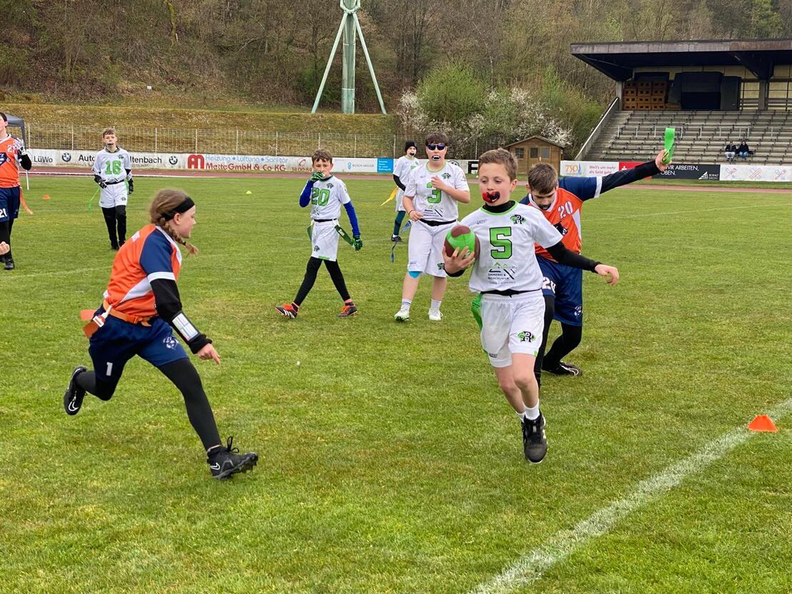 Kinder spielen Football auf einem gr&uuml;nen Rasenplatz, ein Junge l&auml;uft mit dem Ball, w&auml;hrend ein anderer ihn verfolgt.