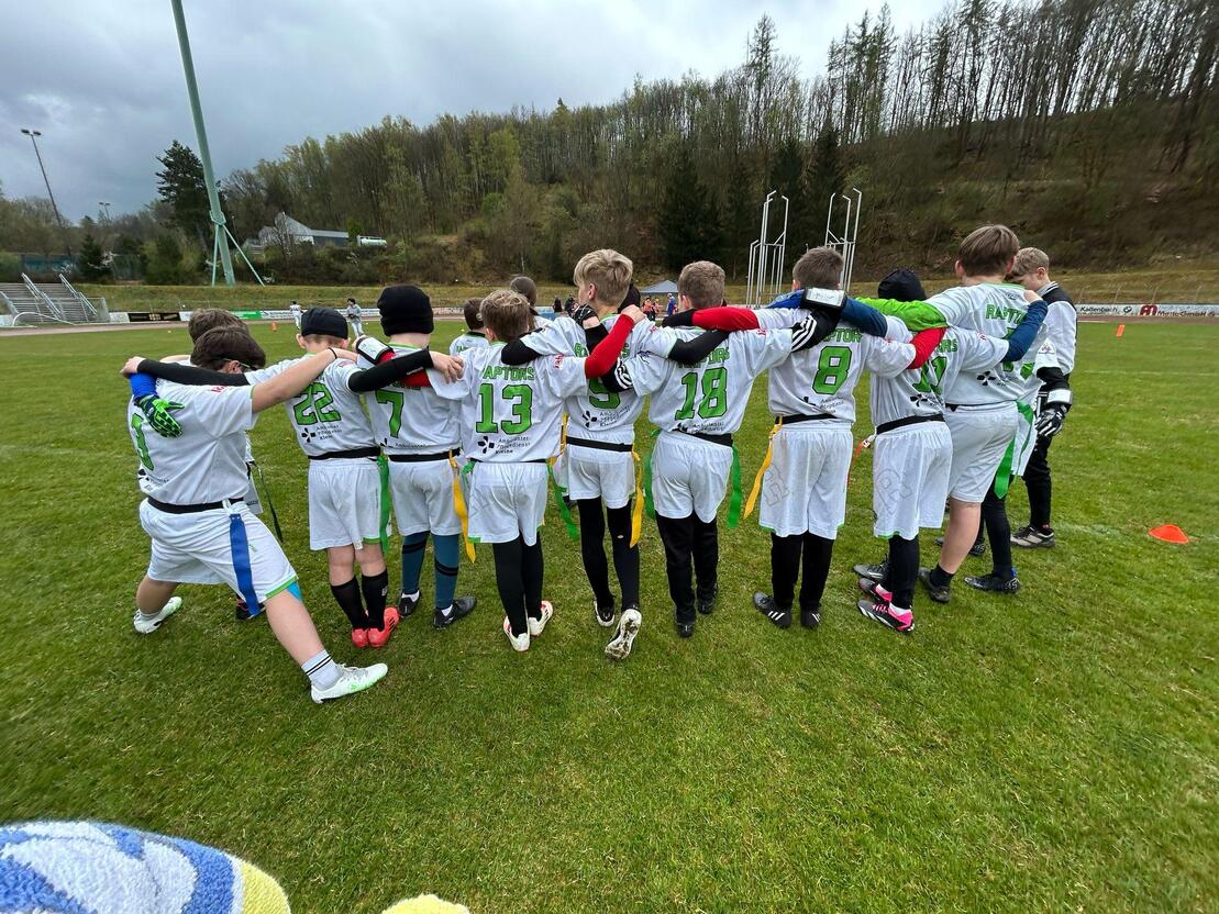 Gruppenspieler in wei&szlig;en Trikots mit Nummern stehen auf einem Sportfeld und umarmen sich, im Hintergrund B&auml;ume und Sportger&auml;te.