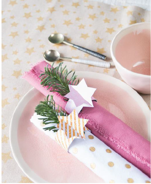 Tischdeko mit pinker Serviette, einer wei&szlig;en Serviette mit Sternen und gr&uuml;nen Zweigen auf einem Teller. Im Hintergrund L&ouml;ffel und Sch&uuml;ssel.