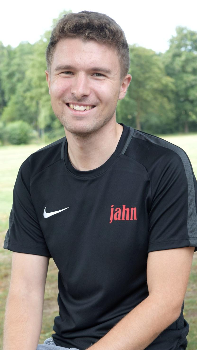 Junger Mann mit kurzem Haar, l&auml;chelnd in schwarzem Shirt, sitzt auf einem Holzstamm vor gr&uuml;nem Hintergrund.