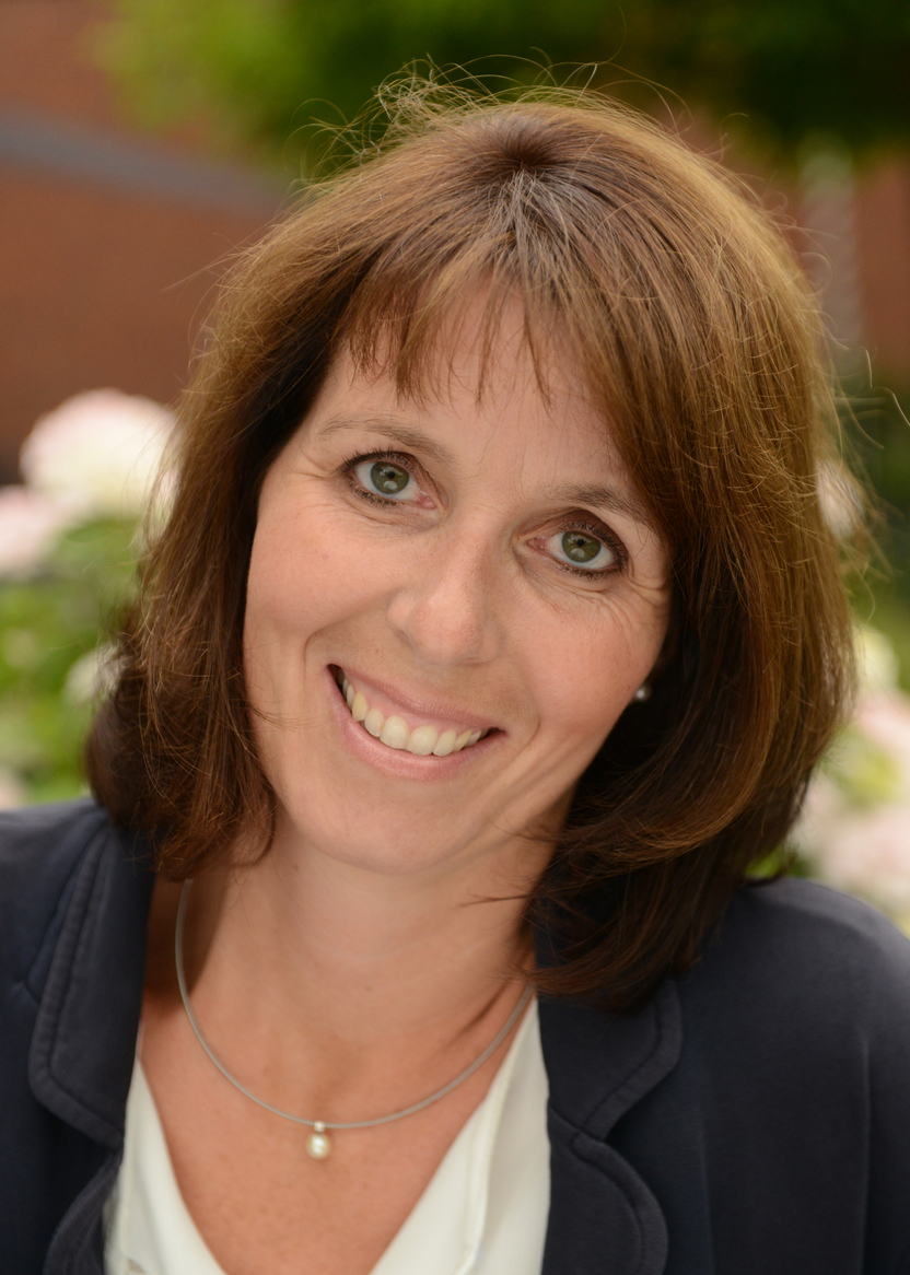 Frau mit braunen Haaren l&auml;chelt vor einer bl&uuml;henden Pflanzenlandschaft, tr&auml;gt ein graues Jackett und eine Perlenkette.