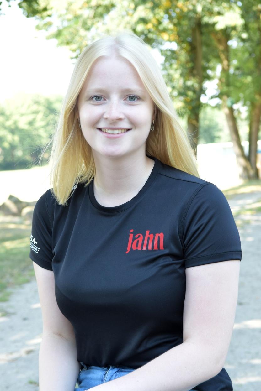 Junge Frau mit blonden Haaren in schwarzem Shirt mit rotem Schriftzug, l&auml;chelt im Freien vor unscharfem Hintergrund.