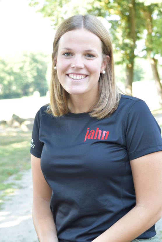 L&auml;chelnde Frau mit kurzen, blonden Haaren in einem schwarzen T-Shirt mit rotem Schriftzug, im Freien vor B&auml;umen.