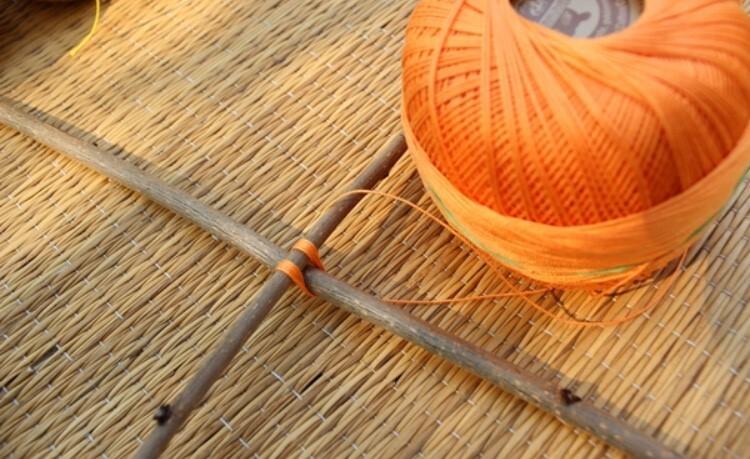 Oranger Faden auf einer Rolle und ein Knoten an einem Holzstab, auf einem geflochtenen Untergrund.