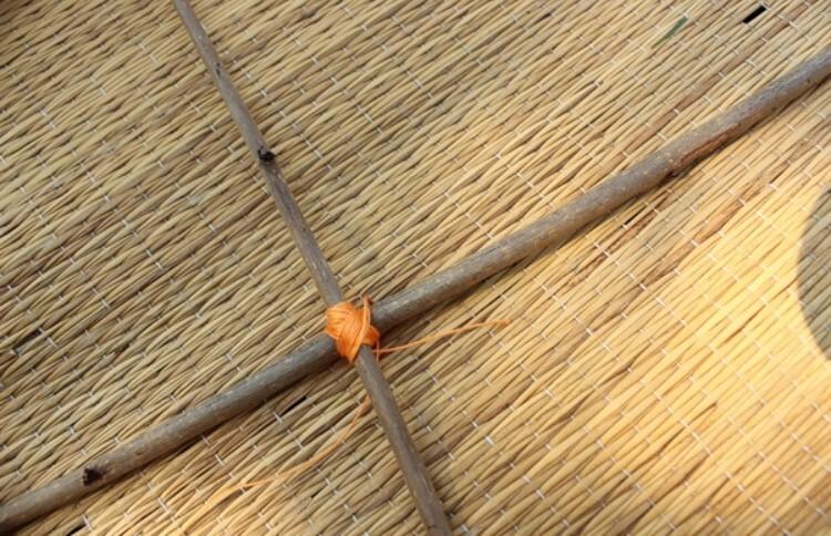 Kreuzung von zwei Holzst&auml;ben, verbunden mit orangefarbenem Band, auf einem matten Untergrund aus geflochtenem Material.