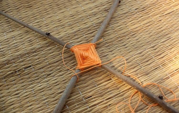 Oranger Faden ist um einen quadratischen Knoten gewickelt, der auf einem geflochtenen Untergrund aus Naturmaterialien liegt.