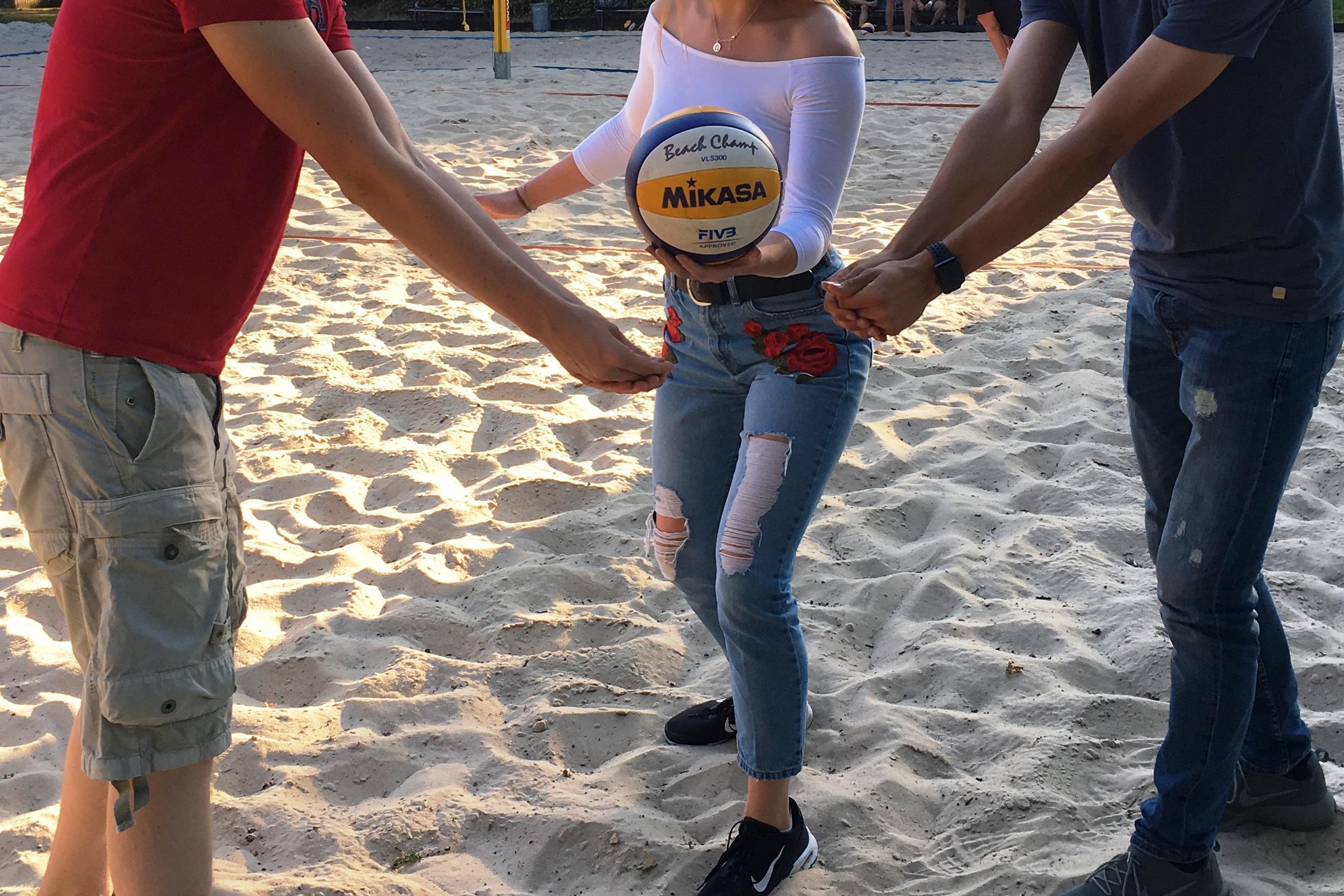 Drei Personen stehen auf einem Sandplatz und halten gemeinsam einen Volleyball in der Hand.